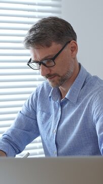 Businessman wearing blue sleek shirt, glasses, is thinking and concentrating while working at the desk in a bright, modern office, demonstrating professionalism and efficiency. Business people concept