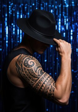 Side View Of Man In Black Tank Top With Tribal Arm Tattoo Tipping Black Fedora Against Bright Blue Sequined Background