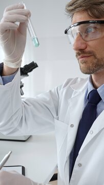 Laboratory professional examining a blue liquid test tube while documenting research in a protective coat, glasses, and gloves. Medicine, health care and science concept