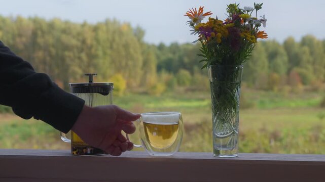 Person takes fresh cup from terrace railing beside glass pot. French press beverage maker and decorative vase overlook scenic autumn fields, creating calm and cozy country morning setting.