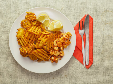Plate holds crispy golden waffle fries and small shrimp. Two lemon wedges add freshness. Red napkin and silverware sit beside plate. Image shows appetizing seafood and fry combo