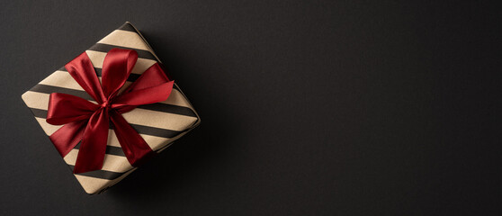 Obraz premium Wrapped gift with red satin bow on banner black wide background for Merry Christmas and Happy New Year