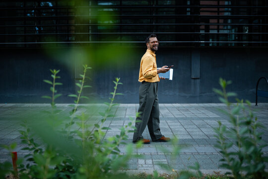 Professional man walking outdoors using phone and laptop