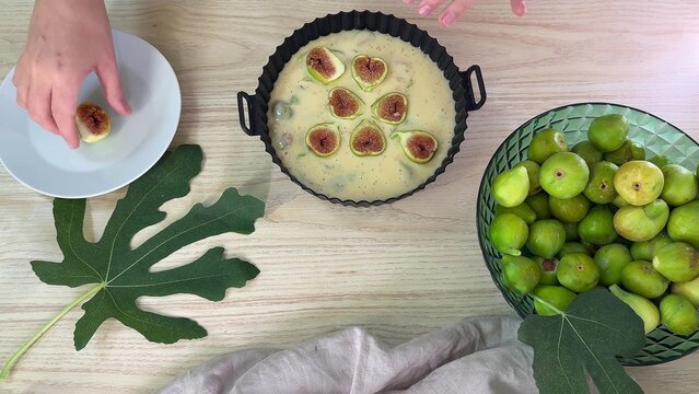 A person handles a sliced fig over a white plate, nearby a black baking pan containing batter topped with fig slices and a bowl of whole green figs on a wooden surface