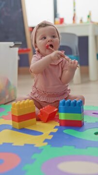 Girl sitting on colorful foam mat, chewing a wooden teether with hands and mouth beside stacking blocks inside building playroom; curiosity exploration.