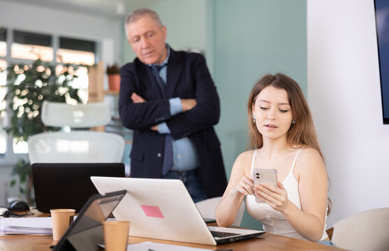 Elderly man sternly scolds young female colleague for using mobile phone while working in office