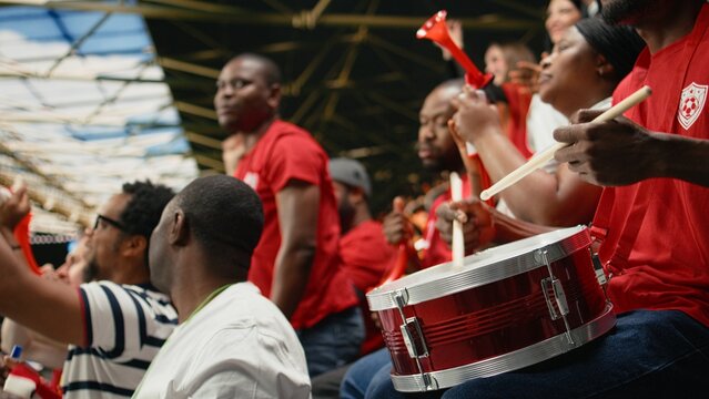 Energetic football supporter beating a percussion drum in the terrace stands, leading chanting fans during live soccer match; hardcore supporters in the fan section celebrate with loud stadium noise.