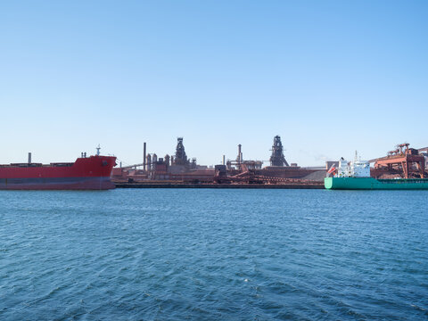 Cargo ship loading steel products at a port near a heavy industrial metallurgical plant. Industrial maritime transportation and environmental impact.