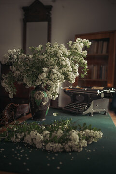 Vintage typewriter and bouquet of white spring flowers in vase on wooden table in moody room.