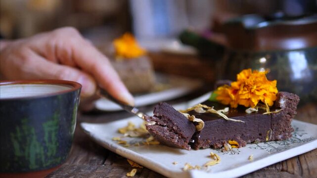 Hand cuts and lifts chocolate tart beside tea cup, preparing dessert for serving. Dense slice breaks away cleanly, with crumbs scattered on plate and warm marigold garnish nearby, ready to eat.