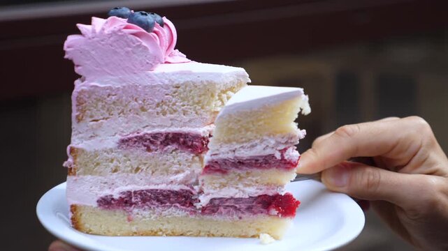 Hand serves layered cake with fork, cutting through creamy berry filling. Dessert slice remains on plate while portion lifts free, showing soft sponge, pink frosting, and fresh blueberry garnish.