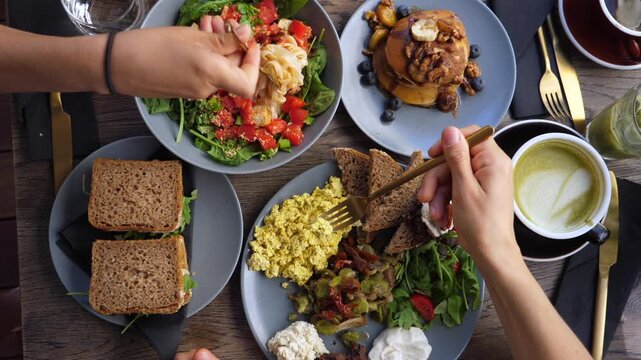 Shared breakfast table fills frame as hands eat from colorful brunch plates with forks. Scrambled eggs, salad, sandwiches, pancakes and coffee suggest leisurely social meal together.