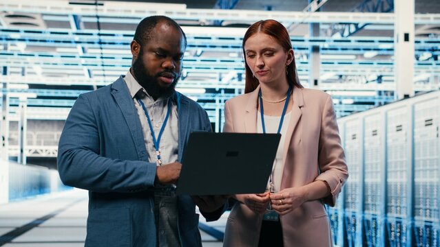 Engineering team in a server hub works together on a laptop to maintain machines while colleagues configure server settings in a high tech data center and complete technical maintenance tasks.