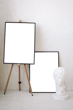 Two blank frame mockups on easel and floor next to plaster bust statue in white minimalist studio art room.