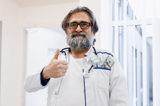 Senior bearded doctor showing thumbs up with money in pocket