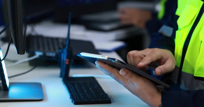 Close-up of a worker in a high visibility jacket using a tablet beside a computer, keyboard and radio in a control room.