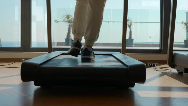 Person walking on a treadmill in a modern gym. Low angle view of a person's feet in sneakers walking on a treadmill in a fitness club in front of a large window with a city and sea view