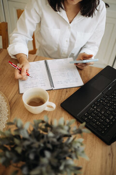Woman writing in notebook with pen holding smartphone near laptop and coffee cup on wooden table.