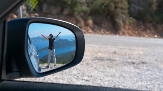 Woman with arms outstretched reflected in car side mirror. Happy woman with her arms raised in freedom is reflected in a car's side mirror, standing on a scenic road with a beautiful sea view