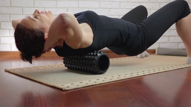 Woman doing foam roller back massage exercise at home. Fit woman in black sportswear performing a myofascial release exercise on a foam roller, massaging her upper back and neck muscles on a yoga mat