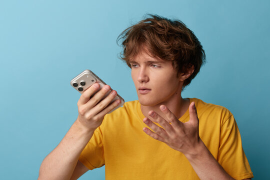 Puzzled young man in yellow t-shirt gestures with open palm while reading shocking news or error on smartphone screen. Blue background, disbelief concept.