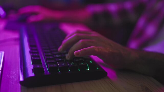 Young man typing on rgb backlit keyboard with hands and fingers on keys, hands resting on wooden desk in studio under purple neon lighting; focus skill play.