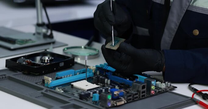 Gloved technician uses a screwdriver to repair a computer motherboard beside a hard drive and tools in an electronics workshop.