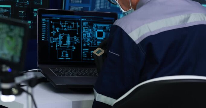 Industrial technician in reflective work wear studies technical diagrams on a laptop in a control room with multiple monitoring screens.