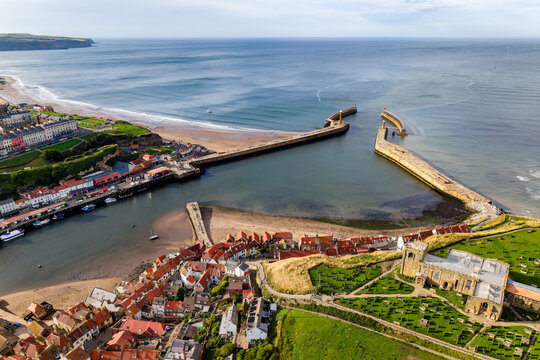 Historic coastal town with twin breakwater piers shielding river mouth from the sea, red pantile rooftops below grassy headland in calm sunny weather