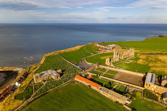 Ruined Gothic abbey and historic stone church on a coastal headland, gravestones across grass fields beside steep cliffs above the sea