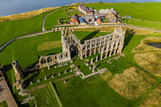 Ruined Gothic abbey church walls and standing pillars on headland grass, North Sea coastline and clifftop farm buildings visible behind the monastery
