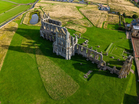 Aerial view of medieval monastery ruins surrounded by green fields, stone arches and tracery casting shadows across grass on sunny day in northern England