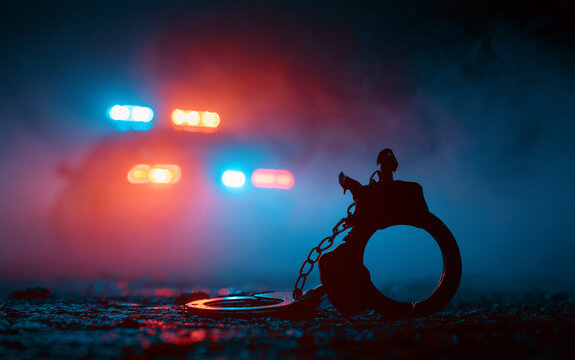 Police raid at night and you are under arrest concept. Silhouette of handcuffs with police car on backside with the flashing red and blue police lights at foggy background
