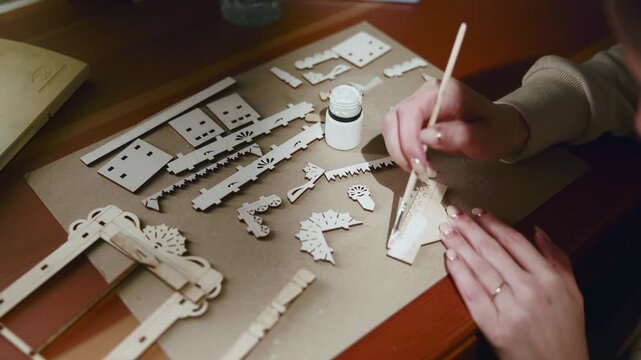 Hands painting laser-cut wooden craft pieces on warm tabletop, showing careful hobby work with tiny house parts, brush, paint jar, and scattered decorative shapes in cozy indoor workspace