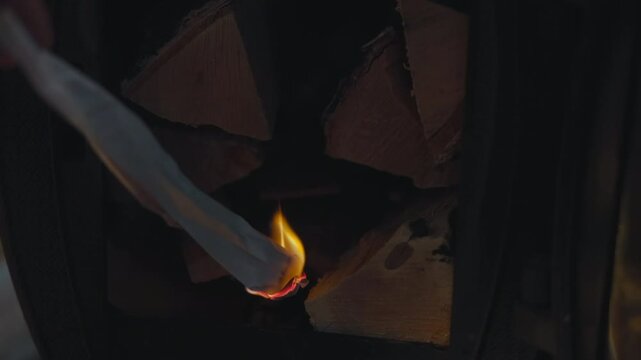 Hand tending fire inside dark fireplace with metal tool, glowing split logs, small flame, charred wood, and warm orange light creating rustic indoor atmosphere with tight dramatic contrast