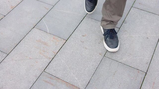 Person walking across gray stone paving in navy sneakers and khaki trousers, shown from downward view with steady step progression, urban texture, soft daylight, and casual everyday mood