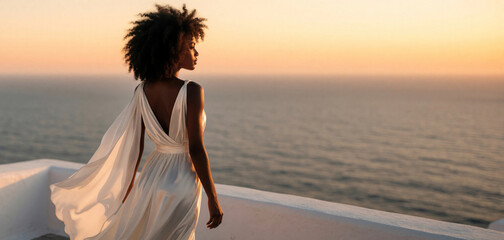 Fototapeta premium Elegant African American woman in white dress stands on Mediterranean terrace balcony. Black female enjoys sunset view over blue sea. Peaceful evening scenery at luxury resort during summer vacation.