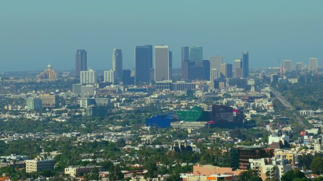 Century City skyscrapers rise above West Hollywood in Los Angeles, California. Telephoto view over palm trees, dense houses and blue, green and red glass buildings on a hazy sunny day. Urban skyline.