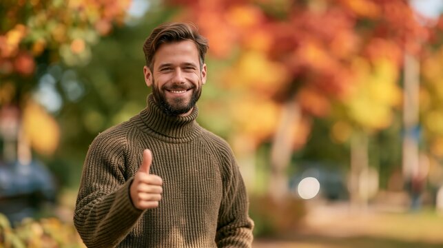 a man standing outdoors in a park during autumn. He wears a textured brown turtleneck sweater and has a beard. The man is smiling and giving a thumbsup gesture with his right hand.