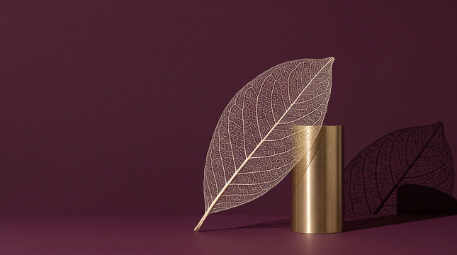 Delicate dried magnolia skeleton leaf leaning on brushed brass cylinder against burgundy backdrop with copy space