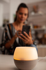 Fototapeta premium Essential oil diffuser glows in sharp foreground focus while a woman uses her smartphone and holds a mug in the background, suggesting calm digital downtime at home, supporting aromatherapy use.