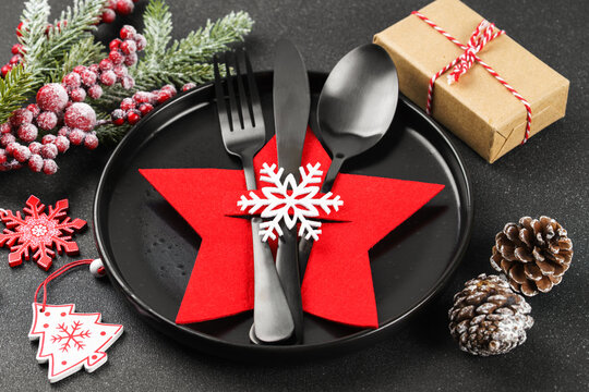 Holiday table setting with black cutlery arranged in red star shaped cutlery holder decorated with white snowflake, Christmas tree branch, ornaments and gift box on dark textured background
