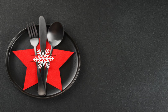 Christmas table setting with black plate and cutlery arranged in red star shaped cutlery holder decorated with white snowflake. Dark textured background with copy space, elegant holiday backdrop.