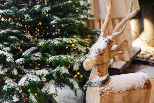 Christmas tree and traditional reindeer decoration made of natural wood beside a snow covered fir tree, with festive lights background and ornaments in a holiday market setting, aesthetic mood
