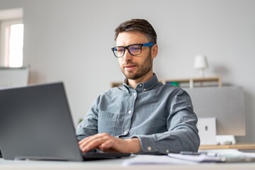 Naklejka na ściany i meble Focused Caucasian Man In His 30s Working On Laptop At A Modern Office Desk, Engaged In Professional Tasks. Creating, Typing, Or Reviewing Documents While Surrounded By Office Equipment.
