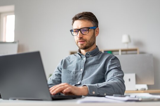 Focused Caucasian Man In His 30s Working On Laptop At A Modern Office Desk, Engaged In Professional Tasks. Creating, Typing, Or Reviewing Documents While Surrounded By Office Equipment.