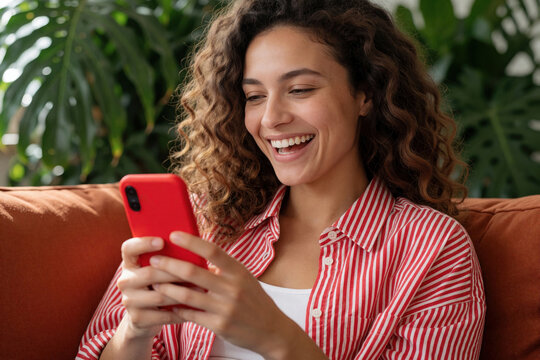 Happy young woman laughs using red smartphone on orange sofa. Joyful girl sits at home looking at mobile screen with big smile. Female user enjoys social media, online chat funny video on cell phone