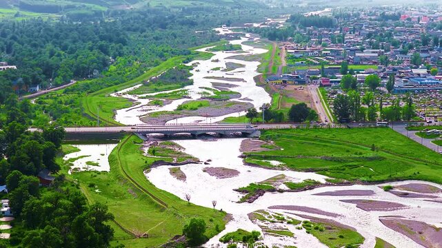 High angle aerial view shows a meandering river flowing through Nalati Grassland town with lush green forests and bridge.