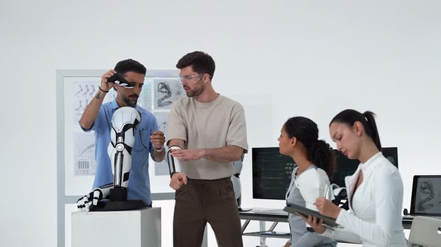 Engineer in blue checks his phone while a colleague in safety glasses examines a wrist brace, then they align a robotic arm beside a robot model as two women sit at monitors