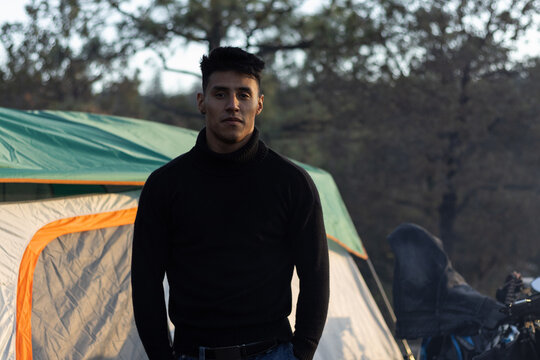 Young man in a black turtleneck sweater standing confidently in front of a camping tent, embracing the outdoor lifestyle, traveling, and experiencing a nature adventure